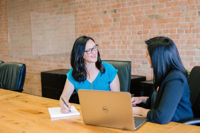 Femme en chemise sarcelle assise à côté d'une autre femme en veste de costume dans un environnement de travail.